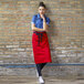 A woman wearing a red Uncommon Chef bistro apron with black webbing standing against a brick wall.