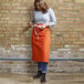 A woman wearing an orange Uncommon Chef bistro apron with natural webbing.