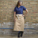 A woman wearing a blue shirt and a light tan Uncommon Chef Marvel Bistro Apron standing in front of a brick wall.