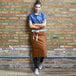 A woman wearing a brown Uncommon Chef Marvel Bistro apron standing in front of a brick wall.