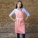 A woman wearing a coral pink Uncommon Chef Vibe apron with natural webbing and three pockets.