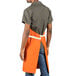 A person wearing an orange Uncommon Chef Vibe bib apron with natural webbing and 3 pockets.