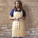 A woman wearing a light beige Uncommon Chef canvas bib apron with natural webbing and 3 pockets.