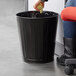 A woman sitting in an office chair throws paper into a black Rubbermaid trash can.