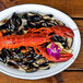 A white enamelware oval platter with a black rolled rim holding a plate of mussels and lobster with a purple flower.