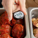 A hand holding a Comark digital pocket probe thermometer with a rubber boot over a tray of meatballs.