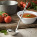 A Carlisle stainless steel ladle in a bowl of tomato soup with a spoon next to a bowl of tomatoes.