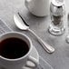 An Acopa stainless steel teaspoon next to a cup of coffee on a table.