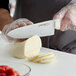 A person using a Dexter-Russell scalloped chef knife to cut a piece of cheese on a table.