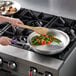 A person cooking vegetables in a Vollrath aluminum fry pan on a stove.