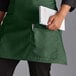 A woman wearing a Choice hunter green half bistro apron with 2 pockets holding a notebook and pen.