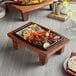 A Valor rectangular cast iron fajita skillet on a rustic chestnut display stand filled with sizzling fajitas on a table.