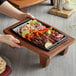 A person holding a Valor rectangular cast iron fajita skillet on a wood tray with meat and vegetables.