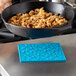 A black pan with food in it being placed on a turquoise Lodge skillet pattern silicone trivet on a table.