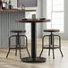A Lancaster Table & Seating cast iron counter height table base with a round black metal top and two stools.