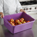 A woman in a chef's uniform holding a Vollrath purple bus tub full of apples.