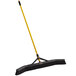 A Rubbermaid commercial push broom with a yellow handle and black bristles.
