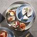 A Corona by GET Enterprises blue porcelain coupe plate with food on it, including a piece of bread with cream cheese and cherry tomatoes.