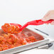 A person holding a red Cambro Camwear salad bar spoon over a tray of food.