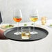 A person holding a Choice black round non-skid serving tray with wine glasses and a glass of ice.