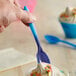 A hand holding a blue and purple dessert spoon over a cupcake.