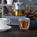 A Villeroy & Boch glass cup of coffee on a table with a teapot.