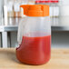 A Tablecraft plastic dispenser jar filled with red liquid and an orange lid.