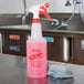 A pink labeled 32 oz. bottle of Noble Chemical All Surf All Purpose Cleaner on a counter with a white cap and a grey cloth.