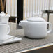 A white teapot with a lid on a table with cups and a plate.