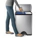 A person in jeans using a simplehuman stainless steel dual compartment recycling bin to throw a plastic bottle in the trash.