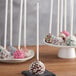 A chocolate covered cake pop with white sprinkles on a white stick.