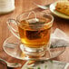 A glass cup of Steep Cafe tea with a tea bag on a saucer.