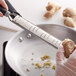 A gloved hand uses a MercerGrates stainless steel shaver to peel ginger into a pan.