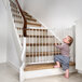 A baby standing next to a white L.A. Baby angle mount safety gate on a staircase.
