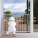 A baby in a white outfit standing next to a white metal extending safety gate.