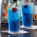 A Duralex highball glass filled with a blue liquid and a cherry with a second glass in the background.