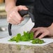 A hand uses a Tablecraft mezzaluna knife to chop mint leaves on a cutting board.