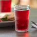 A Duralex glass filled with red liquid on a table with a plate of pancakes.
