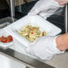 A person in white gloves holding a white Carlisle cold food pan filled with pasta and vegetables.