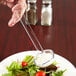 A person using a Thunder Group clear plastic ladle to pour dressing on a plate of salad.