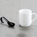 A white mug on a grey surface with a black Fineline Tiny Tensils plastic spoon inside.