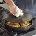 A hand using a white towel to clean a FINEX cast iron cover over food in a pan.
