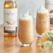 A glass of iced coffee with whipped cream next to a bottle of Capora French vanilla flavoring syrup.