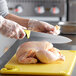 A person using a Dexter-Russell chef knife with a yellow handle to cut a raw chicken on a cutting board.