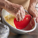 A person's hand using a red silicone bowl scraper to mix dough in a bowl.