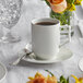 An Acopa bright white porcelain cup filled with a brown liquid on a saucer on a table with a vase of flowers.