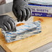A person wearing black gloves using Choice blue striped foil to pack food.