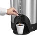 A hand pouring coffee from a Hamilton Beach stainless steel coffee urn into a cup.
