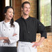 A man and woman wearing Uncommon Chef black 3/4 length chef coats standing next to each other in a professional kitchen.