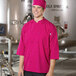 A man in a pink Uncommon Chef 3/4 sleeve chef coat smiles in a professional kitchen.
